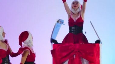 Seksi Kar Kızı elinde bir hediyeyle büyük beyaz bir pastadan çıkıyor. Ayrı bir açık mavi stüdyo arka planında dans eden kırmızı elbiseli genç kadınlar. Kapatın. Yavaş çekim