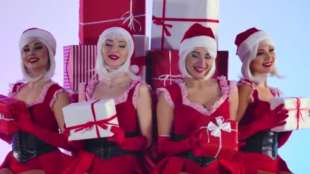 Група молодих сексуальних снігові діви в коротких червоних костюмах і капелюхах сидять з подарунками в руках. Молоді усміхнені жінки позують на тлі подарункових коробок у легкій студії. Близько. Повільний рух . — стокове відео