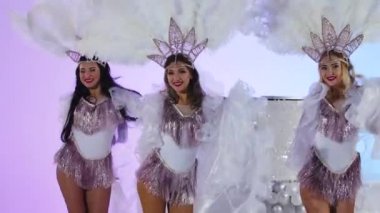 Beyaz Kar Kraliçesi kostümlü ve tüylü başlıklı üç sevimli kadın büyük bir pastanın yanında dans ediyorlar. Dansçılar açık mavi bir stüdyo arka planında poz veriyorlar. Kapatın. Yavaş çekim.