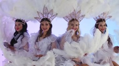 Beyaz Kar Kraliçesi kostümlü ve tüylü başlıklı güzel kadınlar parıldıyor ve eğleniyorlar. Dansçılar açık mavi bir stüdyo arka planında poz veriyorlar. Kapatın. Yavaş çekim.