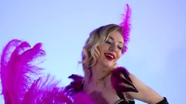 Şampanya bardaklarının yanında siyah bir pastanın üzerinde poz veren üç seksi striptizci. Mavi stüdyo arka planında mor tüyleri olan hoş kadınlar. Kapatın. Yavaş çekim.