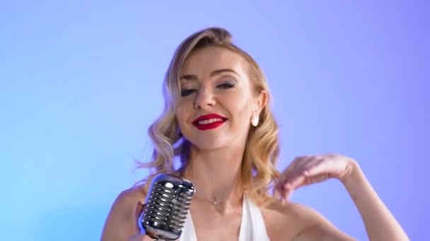Csinos fiatal nő Merlin Monroe képén, amint egy retro mikrofon mellett pózol. Szőke szexuálisan mozog lassított felvételen, világoskék stúdió háttérrel. Közelről.. — Stock videók
