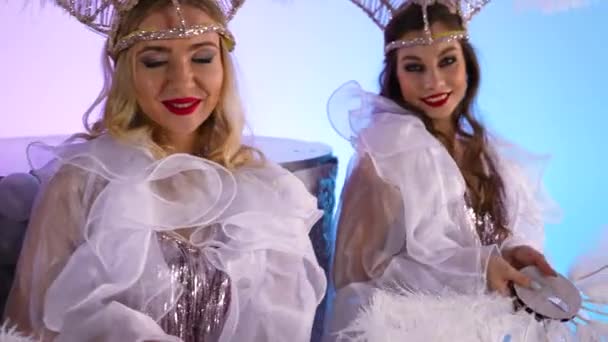 Adorable women in white snow queen costumes and feather headdresses blow glitter and have fun. Dancers posing against a light blue studio background. Close up. Slow motion. — Stock Video