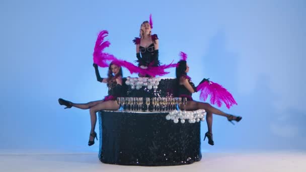 Siyah bir pastanın üzerinde dans eden mor tüylü yelpazeli deri elbiseli seksi genç kadınlar. Stüdyoda açık mavi bir arkaplanda poz veren üç striptizci. Yavaş çekim. — Stok video