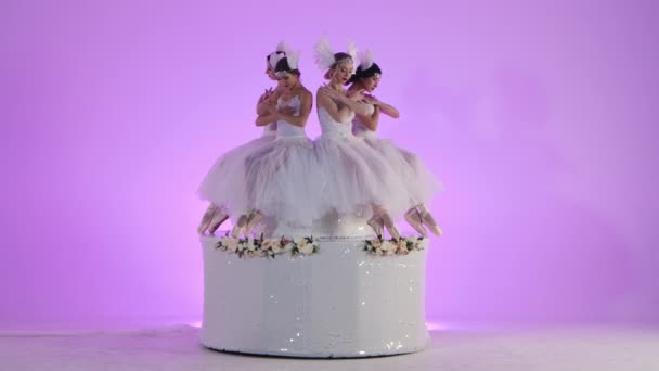 Charmante vrouwelijke ballerina 's gekleed als een witte zwaan dans zittend op de top van een taart versierd met bloemen. Een groep jonge vrouwen beweegt sierlijk in slow motion op roze achtergrond in de studio. — Stockvideo