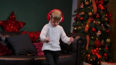 Beyaz kazaklı şirin küçük çocuk büyük kırmızı kulaklıklarla dans ediyor. Parlayan bir Noel ağacının yanındaki süslü bir odada bir genç. Noel arifesi, mutlu çocukluk. Kapatın. Yavaş çekim.