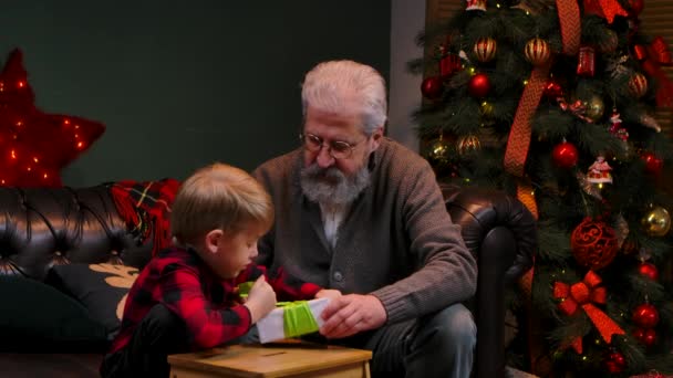 Elegante vecchio e bambino impacchettare confezione regalo e legare un fiocco. Nonno e nipote si siedono su un divano in una stanza decorata vicino a un albero di Natale incandescente. Concetto vacanza in famiglia. Chiudete. Rallentatore. — Video Stock