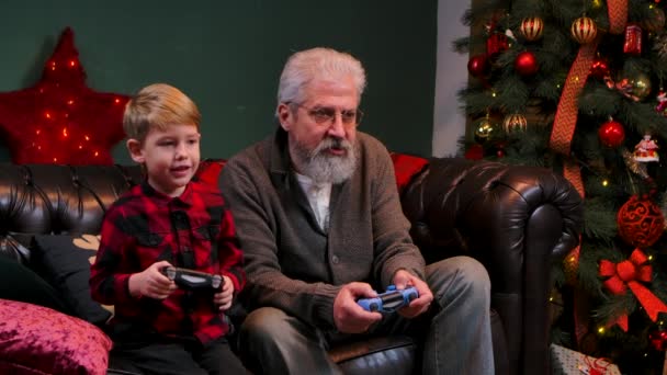 Gelukkige oude grootvader met kleinzoon die een spelletje speelt met joysticks. Elegante grijze man met een jongetje zit op een bank in een versierde kamer bij een gloeiende kerstboom. Sluit maar af. Langzame beweging — Stockvideo