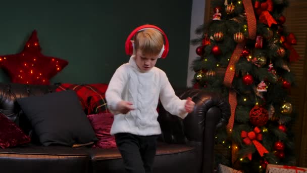 Leuk jongetje in een witte trui danst in een grote rode koptelefoon. Een tiener in een ingerichte kamer in de buurt van een gloeiende kerstboom. Kerstavond, gelukkige jeugd. Sluit maar af. Langzame beweging. — Stockvideo