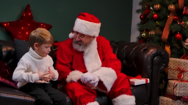 Lindo niño le dice a Santa Claus acerca de sus sueños y recibe un regalo preciado. Nieto y abuelo en un traje rojo y sombrero están sentados en una habitación decorada cerca del árbol de Navidad. De cerca. Movimiento lento. — Vídeos de Stock