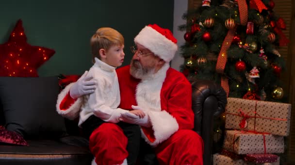 Weihnachtsmann mit kleinem Jungen auf dem Schoß, der über seine Träume spricht. Enkel und Großvater sitzen in einem geschmückten Raum neben einem leuchtenden Weihnachtsbaum und Schachteln mit Geschenken. Aus nächster Nähe. Zeitlupe. — Stockvideo