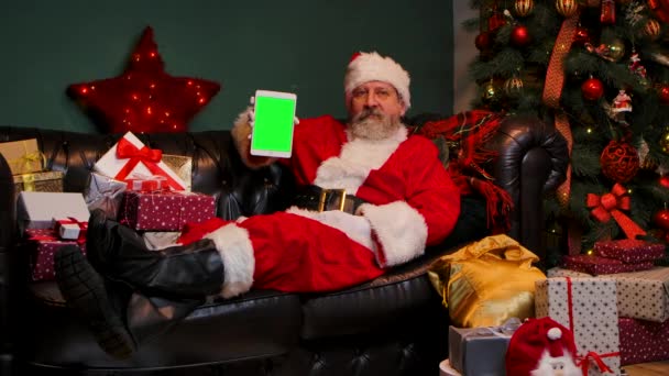 Père Noël se trouve sur le canapé près décoré sapin de Noël et boîtes-cadeaux, montre tablette avec une touche chromée écran vert, faire geste pouce vers le haut. Nouvel An publicité, promotion. Mouvement lent. — Video