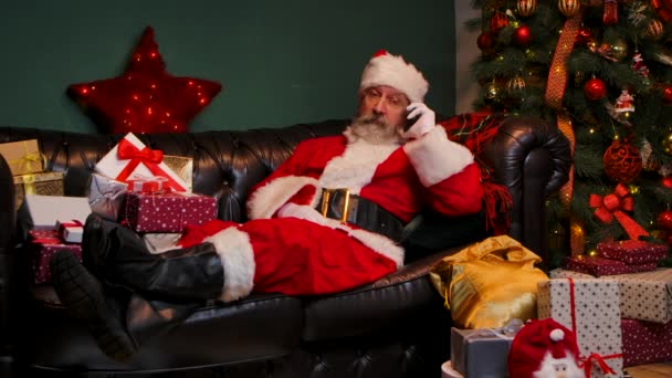 Babbo Natale si trova sul divano vicino a un albero di Natale decorato e scatole regalo, parlando su uno smartphone. Congratulazioni a distanza. Concetto dello spirito di anno nuovo, vacanze e celebrazioni. Rallentatore. — Video Stock
