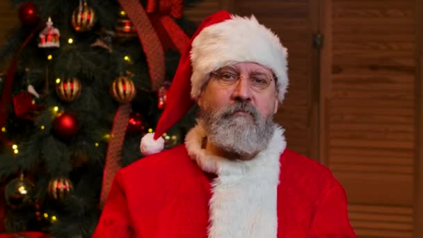Retrato de Santa Claus mira a la cámara en serio, guiña y sonríe. Anciano con barba en traje rojo y sombrero sobre el fondo del árbol de Navidad con juguetes y luces. De cerca. Movimiento lento. — Vídeos de Stock
