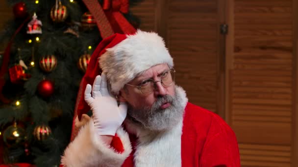 흥미있는 소식에 귀를 기울이려고 손을 가까이 들고 있는 산타클로스의 모습. 빨간 수트에 모자를 쓴 노인이 장난감과 조명을 가지고 크리스마스 트리를 배경으로 하고 있습니다. 닫아. 느린 동작. — 비디오