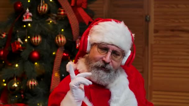 Портрет Санта Клауса, танцующего смешно в больших красных наушниках. Старик с бородой в красном костюме и шляпе на фоне рождественской елки с игрушками и огнями. Закрывай. Медленное движение. — стоковое видео
