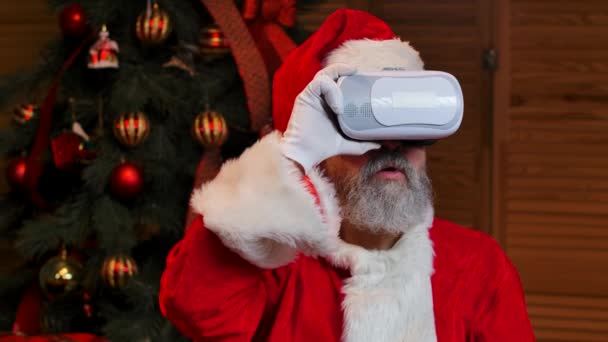 Porträtt jultomten bär virtual reality goggles med wow förvånad uttryck i ansiktet. Gamling med skägg i röd kostym och hatt mot bakgrund av julgran. Närbild. Långsamma rörelser. — Stockvideo
