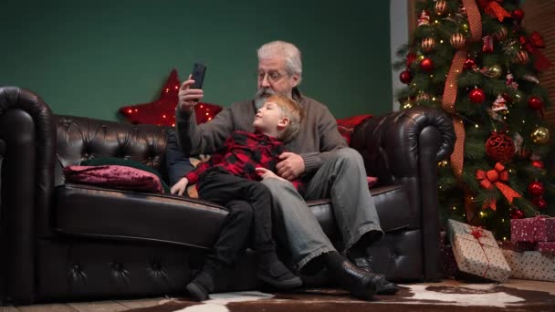 Elegant gammal man och liten pojke talar om video kommunikation på smartphone. Morfar och sonson sitter på en soffa i ett dekorerat rum nära en glödande julgran. Långsamma rörelser. — Stockvideo