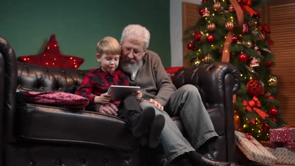 Elegáns öregember és kisfiú beszélgetnek videokommunikáción táblagépen. Nagyapa és unokája ülnek egy kanapén egy díszített szobában, közel egy ragyogó karácsonyfához. Lassú mozgás.. — Stock videók