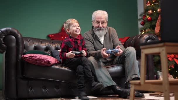 Heureux vieux grand-père avec petit-fils jouer à un jeu avec des joysticks. Élégant homme aux cheveux gris avec un petit garçon est assis sur un canapé dans une pièce décorée près d'un arbre de Noël lumineux. Mouvement lent — Video