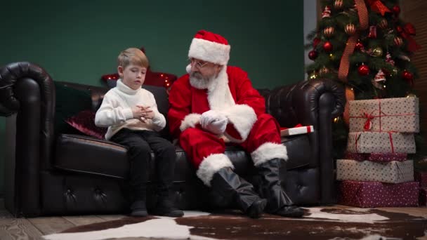 Schattig jongetje vertelt de Kerstman over zijn dromen en ontvangt gekoesterd cadeau. Kleinzoon en grootvader in een rood pak en hoed zitten in een ingerichte kamer in de buurt van de kerstboom. Langzame beweging. — Stockvideo