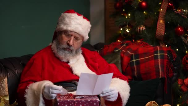 Père Noël lit des lettres et choisit des cadeaux pour les enfants. Vieil homme en costume rouge et un chapeau est assis sur un canapé dans une pièce décorée près d'un arbre de Noël lumineux et des boîtes avec des cadeaux. Ferme là. Mouvement lent. — Video