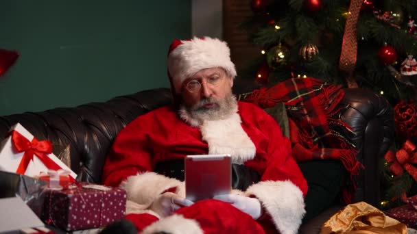 Julemanden kommunikerer via videoopkald ved hjælp af tablet. Gammel mand med skæg i rødt jakkesæt og hat liggende på sofaen i dekoreret værelse nær glødende juletræ og kasser med gaver. Tæt på. Langsom bevægelse. – Stock-video