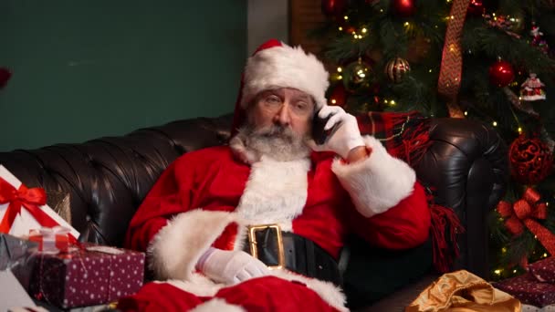 Jultomten ligger på soffan nära en dekorerad julgran och presentaskar, talar på en smartphone. Fjärrgratulationer. Närbild. Långsamma rörelser. — Stockvideo