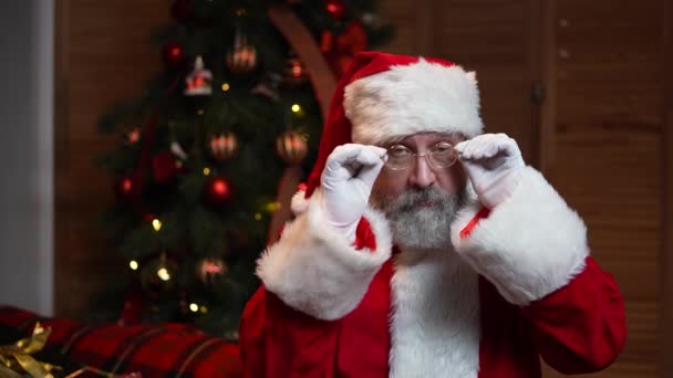 Retrato de Santa Claus mira a la cámara en serio, guiña y sonríe. Anciano con barba en traje rojo y sombrero sobre el fondo del árbol de Navidad con juguetes y luces. Movimiento lento. — Vídeos de Stock