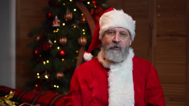Retrato de Santa Claus mirando a la cámara y felicitando a todos en las fiestas. Anciano con barba en traje rojo y sombrero sobre el fondo del árbol de Navidad. Feliz Año Nuevo. Movimiento lento — Vídeos de Stock