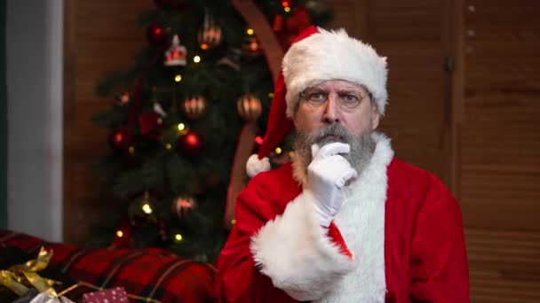 Retrato de Papai Noel olhando penosamente ao redor, faz gesto secreto e pisca. Velho com barba em terno vermelho e chapéu no fundo da árvore de Natal com brinquedos e luzes. Movimento lento. — Vídeo de Stock