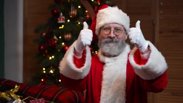 Portrait du Père Noël regardant dans la caméra montrant les pouces vers le haut geste. Vieil homme avec barbe en costume rouge et chapeau sur le fond de l'arbre de Noël avec des jouets et des lumières. . Mouvement lent — Video