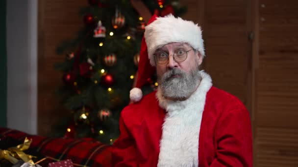 Porträt des Weihnachtsmannes schaut aufmerksam in die Kamera und macht eine Geste in Ordnung. Alter Mann in rotem Anzug und Hut vor dem Hintergrund des Weihnachtsbaums mit Spielzeug und Lichtern. Zeitlupe. — Stockvideo