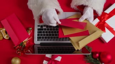 Noel Baba 'nın elleri beyaz eldivenlerle kırmızı yeni yıl süsleme masasında klavye bilgisayarında daktilo ediyor. Noel Baba çocuklardan gelen mektuplara bakar ve internetten hediyeler alır. Kapatın. Yavaş çekim.