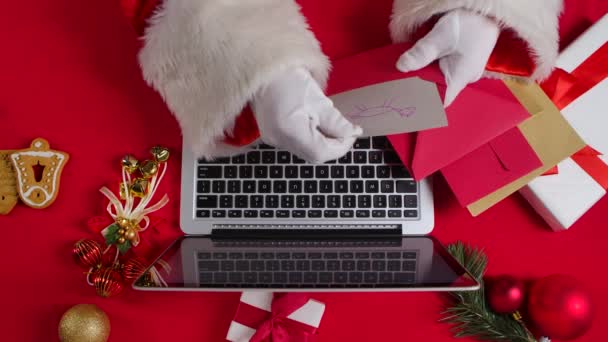 Vista superior Las manos de Santa en guantes blancos están escribiendo en el ordenador portátil del teclado por la mesa roja decorada Año Nuevo. Santa Claus mira a través de cartas de los niños y compra regalos en Internet. De cerca. Movimiento lento. — Vídeo de stock
