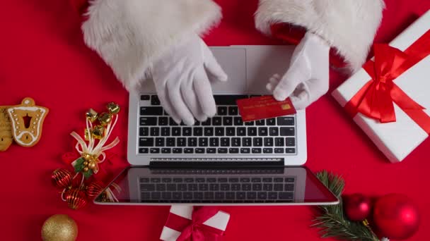 Top view Santa hands in white gloves are typing on the keyboard by red New Year decorated table. Santa Claus pracuje s notebookem při nakupování online s kreditní kartou. Zavřít. Zpomalený pohyb. — Stock video