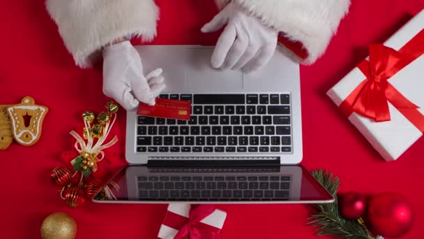 Top view Santa hands in white gloves are typing on the keyboard by red New Year decorated table. Santa Claus works with a laptop while shopping online with a credit card. Close up. Slow motion. — Stock Video