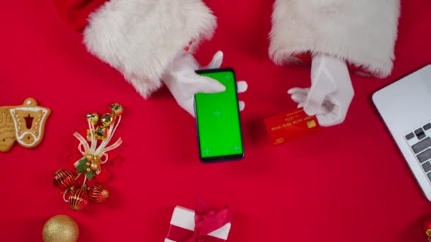 Vue du dessus Les mains du Père Noël en gants blancs utilisent un smartphone avec une clé chromatique à écran vert par une table rouge décorée du Nouvel An. Père Noël fait des achats en ligne avec carte de crédit et téléphone. Ferme là. Mouvement lent. — Video