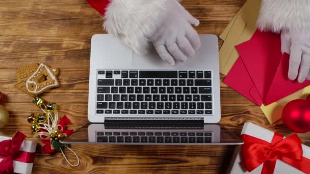 Top view Santa hands in white gloves are typing on keyboard laptop by wooden New Year decorated table. Santa Claus prohlíží dopisy od dětí a nakupuje dárky na internetu. Zavřít. Zpomalený pohyb — Stock video