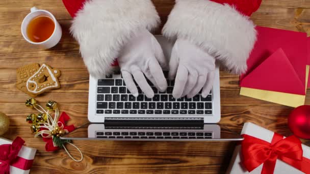 Widok z góry Mikołaj ręce są wpisując na klawiaturze, napoje herbata przez drewniany stół urządzone. Święty Mikołaj pracuje z laptopem, przegląda listy od dzieci i kupuje prezenty w Internecie. Zamknij drzwi. Zwolniony ruch. — Wideo stockowe