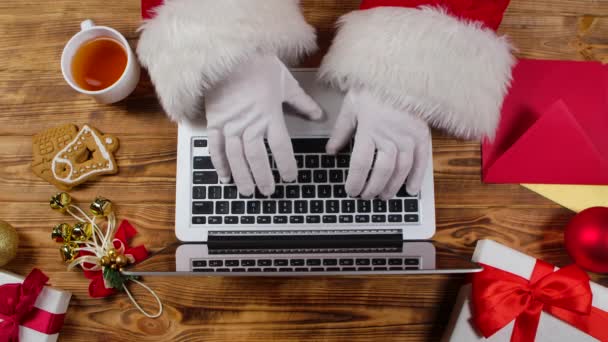 Pohled shora Santa hands are typing on keyboard, drinky čaj by wooden decorated table. Santa Claus pracuje s laptopem, prohlíží si dopisy od dětí a nakupuje dárky na internetu. Zavřít. Zpomalený pohyb. — Stock video