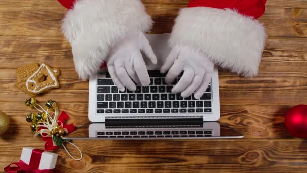 Top view Santa hands in white gloves typing on the keyboard by wooden New Year decorated table. Santa Claus pracuje s laptopem, prohlíží poštu a připravuje dárky pro děti. Zavřít. Zpomalený pohyb — Stock video