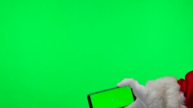 Noel Baba beyaz eldivenler içinde yeşil ekran krom anahtarı olan bir akıllı telefonu yatay pozisyonda tutar. Stüdyoda yeşil bir arka planda izole edilmiş. Ekranı düzelt. Kapatın. Yavaş çekim.
