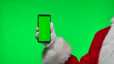Noel Baba beyaz eldivenler veriyor. Akıllı telefonu tut ve yeşil ekran krom anahtarını işaret et. Stüdyoda yeşil bir arka planda izole edilmiş. Ekranı düzelt. Kapatın. Yavaş çekim.