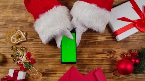 顶部视图圣诞老人的手使用的智能手机与绿色屏幕彩色键木新年装饰桌子。圣诞老人拍打着屏幕看内容，在网上购物。靠近点慢动作. — 图库视频影像