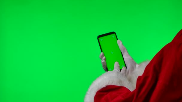 Les mains du Père Noël dans des gants blancs à l'aide d'un smartphone, faites glisser et tapotez sur la touche chromatique de l'écran vert. Isolé sur fond vert dans le studio. Affichage maquillé. Ferme là. Mouvement lent. — Video