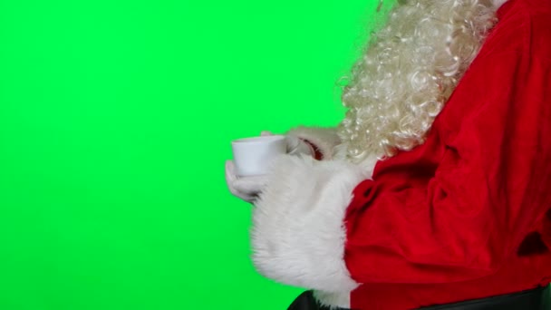 Бічний вид на Санта-Клауса з бородою в білих рукавицях і червоним костюмом зігріває руки на чашці і полюбляє гарячий чай. Розташований на зеленому екрані в студії. Ключ від хроми. Закрийся. Повільний рух. — стокове відео