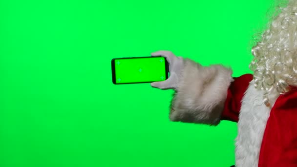 Vista lateral Papai Noel com uma barba em luvas brancas segure um smartphone em uma posição horizontal com uma chave de croma de tela verde. Isolado em um fundo verde no estúdio. Fecha. Movimento lento — Vídeo de Stock