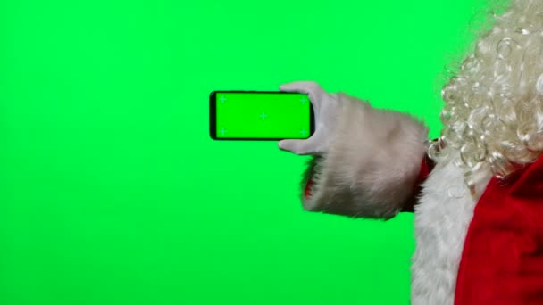 Kerstman met baard in witte handschoenen houdt een smartphone in een horizontale positie en wijst met zijn vinger met groene scherm chroma sleutel. Geïsoleerd op groene achtergrond in de studio. Sluit maar af. Langzame beweging. — Stockvideo