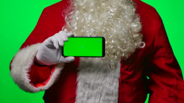 Le Père Noël avec la barbe en gants blancs tient un smartphone dans une position horizontale et pointe son doigt avec la touche chromatique de l'écran vert. Isolé sur fond vert dans le studio. Ferme là. Mouvement lent. — Video